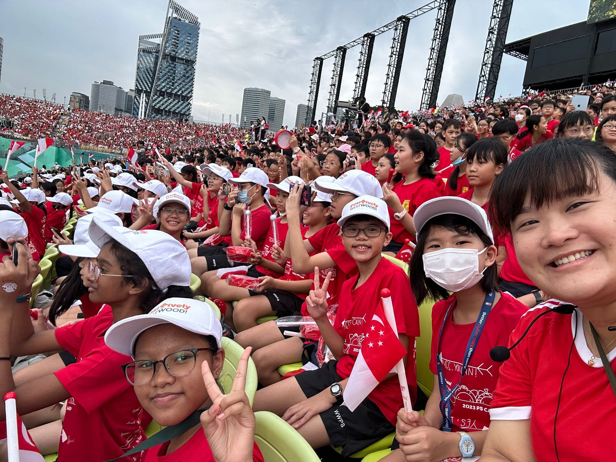 Large crowd of children in red shirts and white caps waving Singapore flags at an outdoor National Day event.